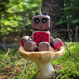 Puede incluir: Una figura de mariquita de madera con detalles rojos y negros se encuentra sobre una seta en un entorno forestal. La mariquita tiene una cara sonriente y está hecha de bloques de madera geométricos. La seta tiene un sombrero beige y un tallo blanco.
