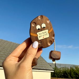 May include: A wooden potato-shaped ornament with painted eyes and a small sign that reads "You have got this!!" with a heart. The ornament is held by a hand against a blue sky background. A small wooden cube hangs from the ornament.