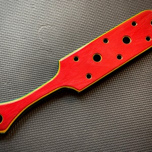 May include: A red wooden paddle with a green and yellow trim. The paddle has multiple round holes cut out of the surface.