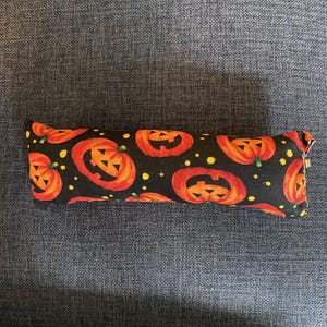 May include: A rectangular fabric item with a Halloween pumpkin pattern. The fabric is black with orange jack-o'-lanterns and small yellow dots. The item is resting on a dark gray surface.