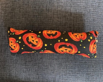 May include: A rectangular fabric item with a Halloween pumpkin pattern. The fabric is black with orange jack-o'-lanterns and small yellow dots. The item is resting on a dark gray surface.