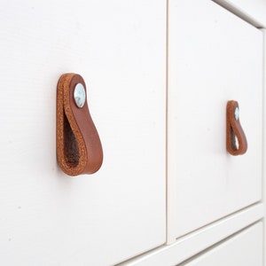 Könnte beinhalten: Nahaufnahme eines weißen Schranks mit braunen Lederschlaufen als Griffen. Die Griffe sind mit silberfarbener Hardware befestigt. Die Schranktüren sind sauber und modern. Für Wohn- und Stauraum.