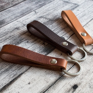 Könnte beinhalten: Drei Leder-Schlüsselanhänger in Braun-, Dunkelbraun- und Beigetönen. Jeder Schlüsselanhänger hat eine Lederschlaufe, die mit einem silberfarbenen Metallring mit einer Metallniete verbunden ist. Die Schlüsselanhänger sind auf einer hellen Holzoberfläche angeordnet.