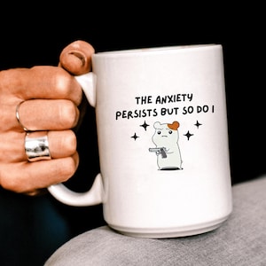 May include: White ceramic mug with a black and white illustration of a hamster holding a gun. The text on the mug reads "The anxiety persists but so do I."