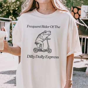 Może przedstawiać: Biały t-shirt z grafiką przedstawiającą żabę jadącą na skuterze. Tekst na koszulce brzmi "Frequent Rider Of The Dilly Dally Express".