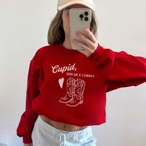 Pode incluir: Camisola vermelha com um gráfico branco que diz "Cupido, Envie-me um Cowboy" e um desenho de duas botas de cowboy.