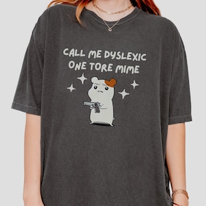 May include: A dark gray t-shirt with the text "Call me dyslexic one tore mime" and a cartoon hamster holding a gun. The hamster is white with brown ears and a black nose.