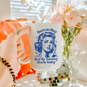 May include: White ceramic mug with a blue illustration of a woman with a tear on her cheek. The text on the mug reads "Born to Slay But My Tummy Hurts Today."
