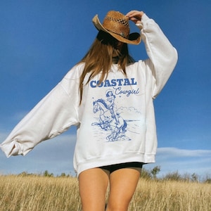 Könnte beinhalten: Weißer Pullover mit einer blauen Grafik einer Frau, die ein Pferd reitet. Die Grafik lautet "Coastal Cowgirl".