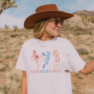 Puede incluir: Camiseta blanca con un gráfico de tres mujeres con sombreros de vaquero leyendo libros. El texto "LET'S READ GIRLS" está impreso debajo del gráfico.