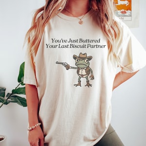 May include: Cream-colored t-shirt featuring the text "You've Just Buttered Your Last Biscuit Partner" above a cartoon frog dressed as a cowboy, holding a gun. The frog is wearing a brown hat, vest, and boots. The shirt is a relaxed fit.