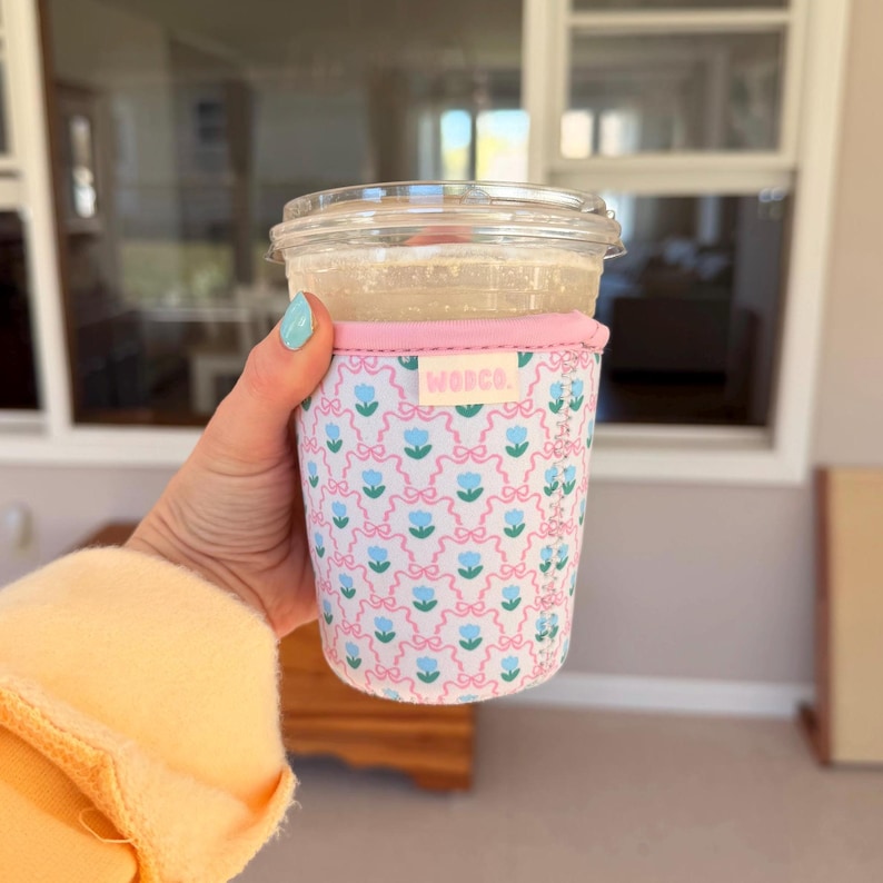 May include: A clear plastic cup containing a drink, held in a hand. The cup is wrapped in a pink and white patterned sleeve featuring blue flowers and bows. The sleeve has the word "WOPCO." printed on it.