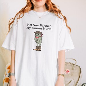 May include: White t-shirt featuring the text "Not Now Partner My Tummy Hurts" above a cartoon frog wearing a cowboy hat, red bandana, and boots. The frog is clutching its stomach. The shirt is a casual, comfortable style.
