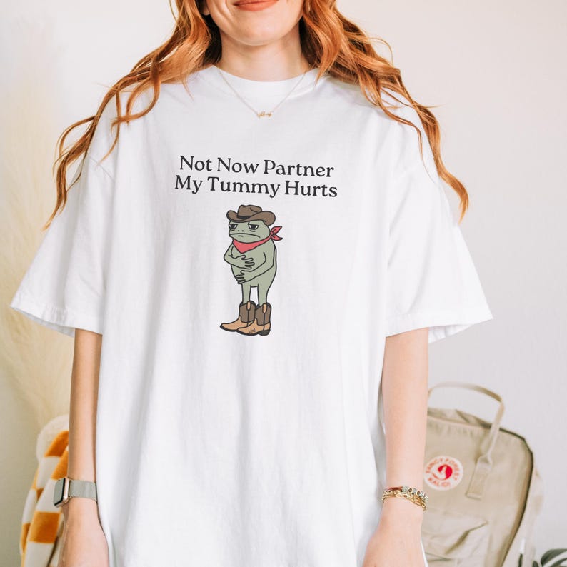 May include: White t-shirt featuring the text "Not Now Partner My Tummy Hurts" above a cartoon frog wearing a cowboy hat, red bandana, and boots. The frog is clutching its stomach. The shirt is a casual, comfortable style.