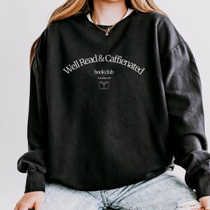 Puede incluir: Sudadera negra con el texto "Well Read & Caffeinated bookclub Established 2017" en letras blancas.
