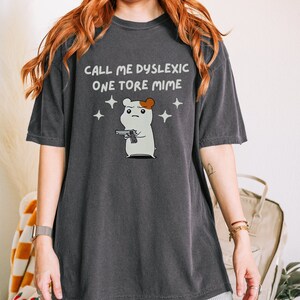 May include: A dark gray t-shirt with the text "CALL ME DYSLEXIC ONE TORE MIME" and a cartoon hamster holding a gun.