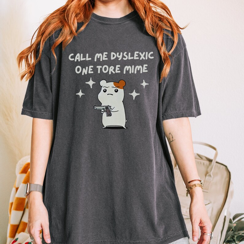 May include: A dark gray t-shirt with the text "CALL ME DYSLEXIC ONE TORE MIME" and a cartoon hamster holding a gun.