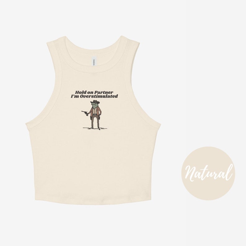 May include: Cream-colored tank top with a humorous graphic of a frog dressed as a cowboy, holding a gun. The text "Hold on Partner I'm Overstimulated" is printed above the frog. The word "Natural" is written in a circle on the right.