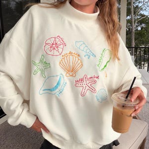 May include: A cream-colored mock neck sweatshirt featuring colorful embroidered sea life motifs including starfish, seashells, and sand dollars. The embroidery includes text labels such as patience, kindness, joy, faithfulness, love, goodness, and self-control.