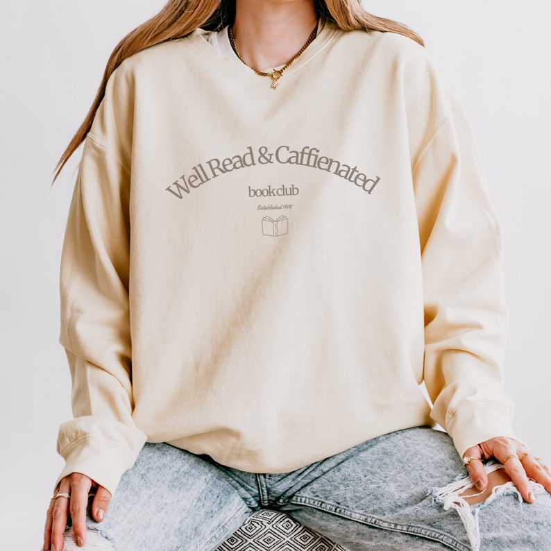 Puede incluir: Una sudadera de color amarillo claro con el texto "Well Read & Caffeinated book club Established 2022" y una peque&ntilde;a imagen de un libro abierto.