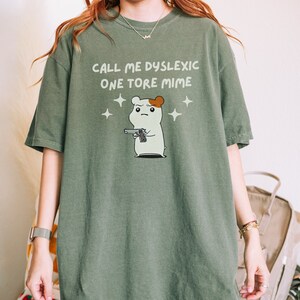 May include: Green oversized t-shirt with a cartoon hamster holding a gun and the text "Call me dyslexic one tore mime".