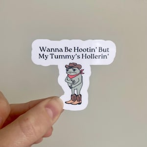 Puede incluir: Una pegatina blanca con una rana de dibujos animados que lleva un sombrero de vaquero y botas. El texto de la pegatina dice "Wanna Be Hootin' But My Tummy's Hollerin'".