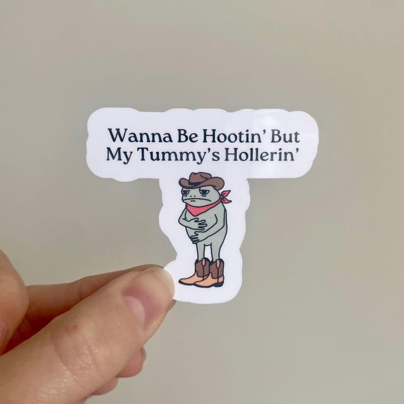 K&ouml;nnte beinhalten: Ein wei&szlig;er Aufkleber mit einem Cartoon-Frosch, der einen Cowboyhut und Stiefel tr&auml;gt. Der Text auf dem Aufkleber lautet "Wanna Be Hootin' But My Tummy's Hollerin'".
