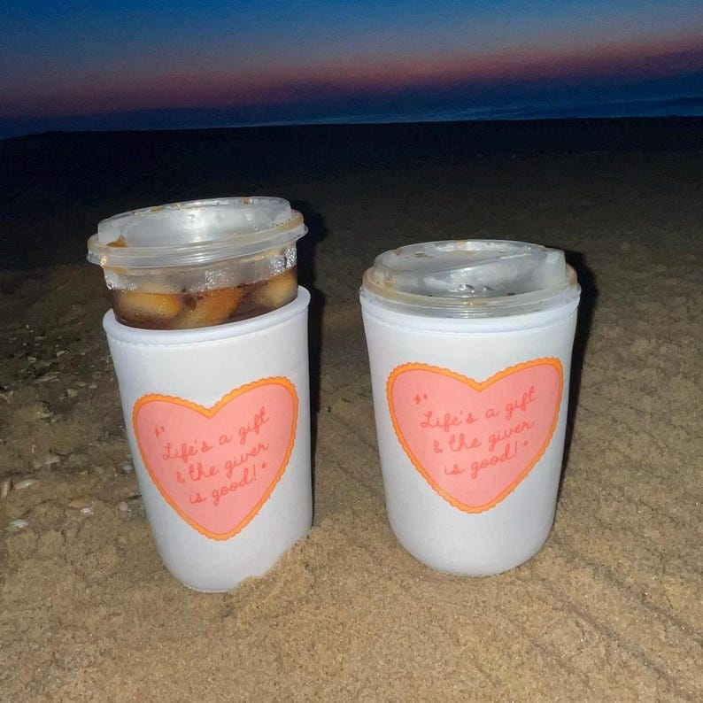 May include: Two white coffee cup sleeves with a pink heart design and the text "Life's a gift & the giver is good!" on a sandy beach. One sleeve holds a clear plastic cup with a beverage.