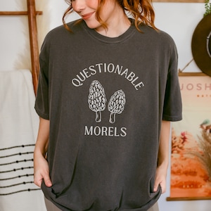 Puede incluir: Una camiseta gris oscuro con un gráfico blanco de dos setas morchella y el texto "Questionable Morels".