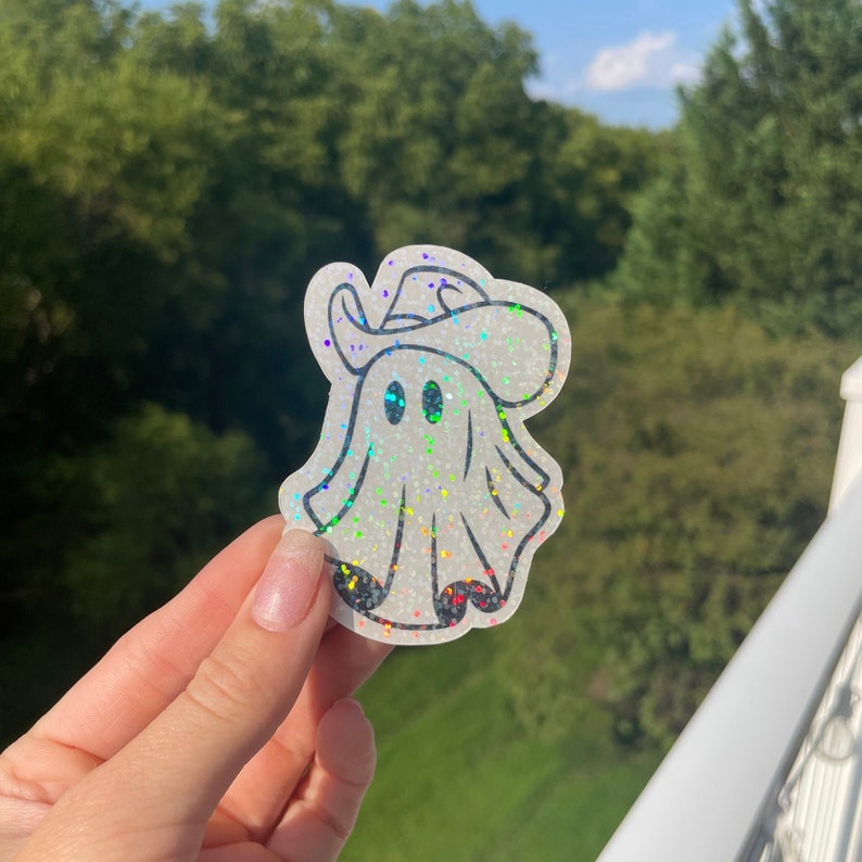 May include: A white iridescent glitter ghost wearing a cowboy hat. The ghost has two black eyes and a white outline.