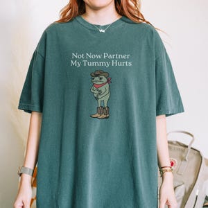 May include: Teal-colored t-shirt featuring a cartoon frog wearing a cowboy hat, boots, and a red bandana. The text "Not Now Partner My Tummy Hurts" is printed above the frog. The frog is holding its stomach.