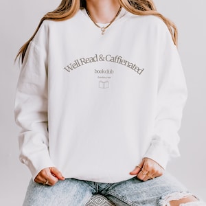 Puede incluir: Sudadera blanca con el texto "Well Read & Caffeinated book club Established 2022" y una peque&ntilde;a imagen de un libro abierto.