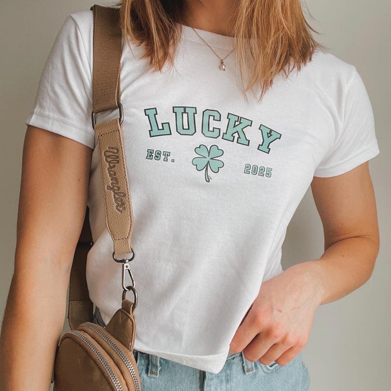 K&ouml;nnte beinhalten: Wei&szlig;es T-Shirt mit einem gr&uuml;nen vierbl&auml;ttrigen Kleeblatt und dem Text "LUCKY EST. 2025" in Blau. Das Shirt wird von einer Person mit einer braunen Leder-Umh&auml;ngetasche mit dem Wort "Wrangler" auf dem Riemen getragen.