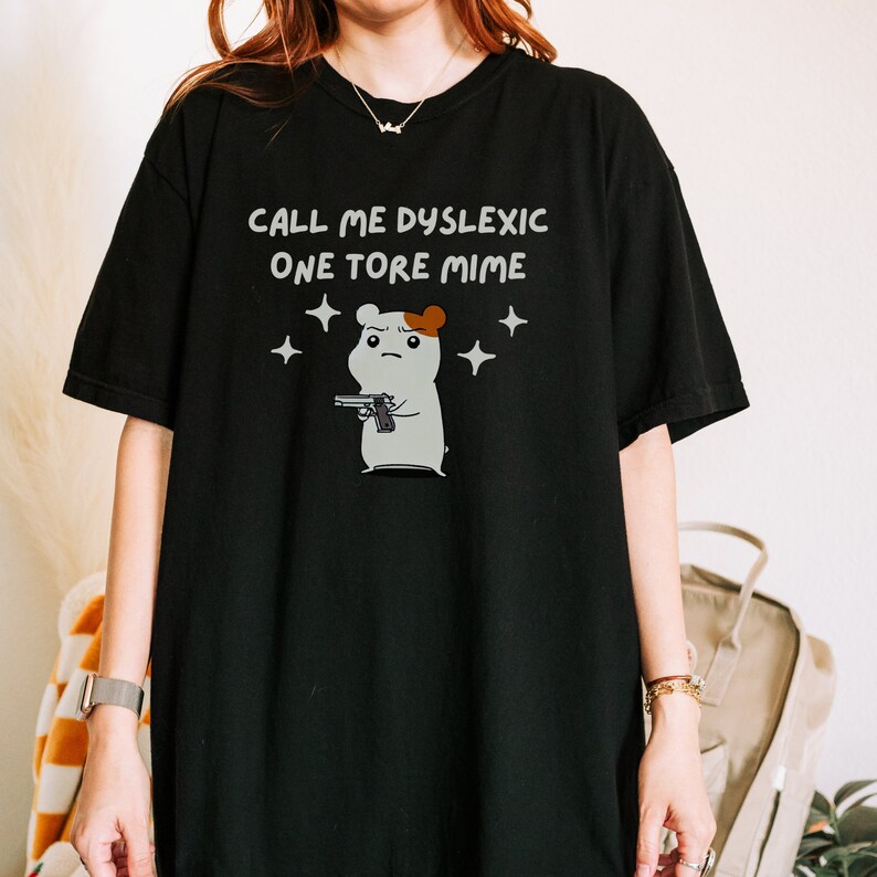 May include: Black t-shirt with a white graphic of a cartoon hamster holding a gun. The text "CALL ME DYSLEXIC ONE TORE MIME" is printed on the shirt.