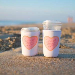 May include: Two white drink sleeves with a pink heart design and the text "Life's a gift & the giver is good!" on a sandy beach. The sleeves are around clear glass cups with white foam.