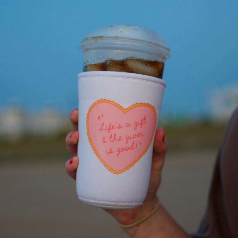 May include: A white coffee cup sleeve with a pink heart design. The heart contains the text "Life's a gift & the giver is good!" in cursive. The sleeve is wrapped around a clear plastic cup filled with iced coffee.