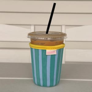 May include: A clear plastic iced coffee cup with a black straw inside a teal and light blue striped fabric sleeve. The sleeve features a bright yellow top trim and a small white rectangular tag with the pink text WODCO. printed on the front.