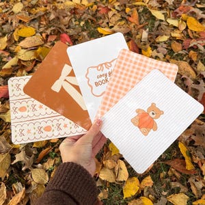 May include: A hand holds a collection of square paper cards with various designs. One card features a bow, another says "cozy gi BOOK", and another has a teddy bear. The cards are in shades of orange, white, and brown.