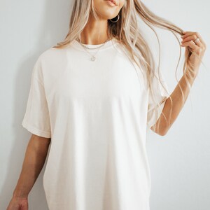 May include: Cream-colored, short-sleeved t-shirt. The shirt has a relaxed fit and a crew neck. The fabric appears to be soft and slightly textured. The model is wearing a silver necklace and earrings. The background is a plain white wall.