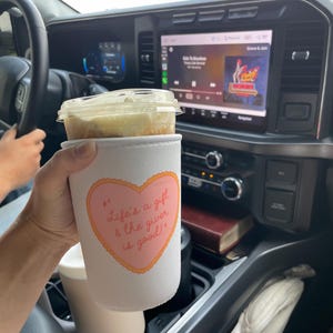 May include: A white coffee cup sleeve with a pink heart design and the text "Life's a gift & the giver is good!" is held in a car. The cup contains a beverage with a clear lid. The car's dashboard and steering wheel are visible.