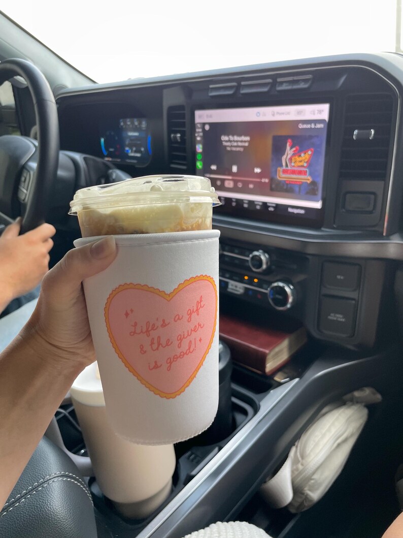 May include: A white coffee cup sleeve with a pink heart design and the text "Life's a gift & the giver is good!" is held in a car. The cup contains a beverage with a clear lid. The car's dashboard and steering wheel are visible.