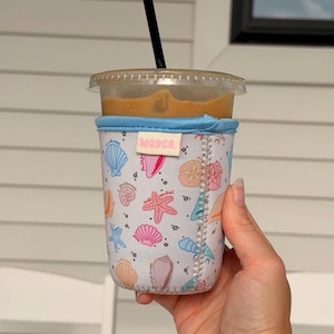 Puede incluir: Una funda de neopreno gris claro para vaso de café helado con un patrón colorido de conchas, estrellas de mar y dólares de arena en tonos rosa, naranja y azul. Una pequeña etiqueta rectangular con el texto WODCO está centrada arriba.