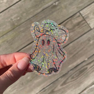 May include: A clear iridescent glitter ghost sticker with a black outline. The ghost is wearing a hat and has two black eyes.