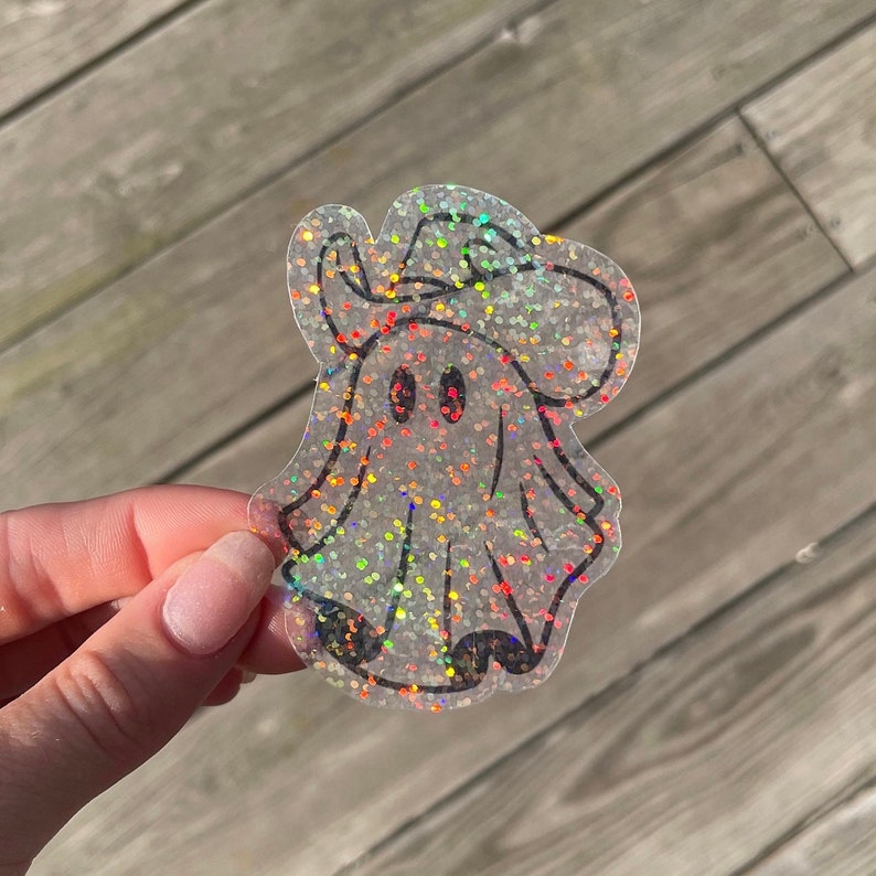 May include: A clear iridescent glitter ghost sticker with a black outline. The ghost is wearing a hat and has two black eyes.