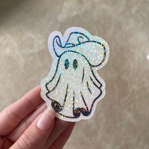 May include: A white iridescent glitter ghost wearing a black cowboy hat with a black outline. The ghost has two black eyes.