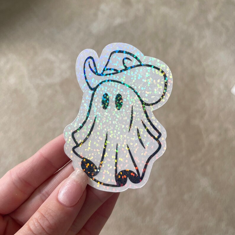 May include: A white iridescent glitter ghost wearing a black cowboy hat with a black outline. The ghost has two black eyes.
