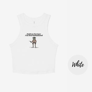 May include: White tank top with a humorous graphic of a frog dressed as a cowboy, holding a gun. The text "Hold on Partner I'm Overstimulated" is above the frog. A white circle with the word "White" in script is on the right.