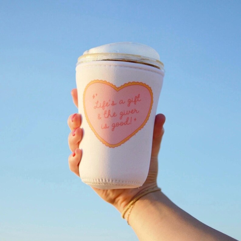 May include: A white insulated beverage holder with a pink heart-shaped design. The heart contains the text "Life's a gift & the giver is good!" in a script font. The holder is held up against a blue sky.