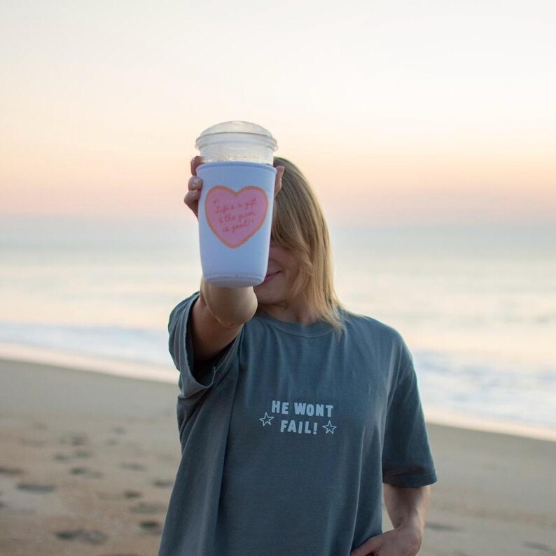 May include: A light blue drink sleeve with a pink heart design and the text "Life is a gift, the giver is good!" is held up in front of a person. The person is wearing a gray t-shirt with the text "HE WONT FAIL!" and two stars.