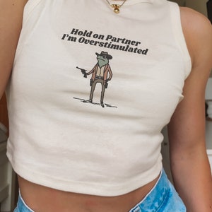May include: Cream-colored tank top with the text "Hold on Partner I'm Overstimulated" and a cartoon frog wearing a cowboy hat and holding a gun. The frog is dressed in a brown jacket and pants. The tank top is paired with blue denim jeans.